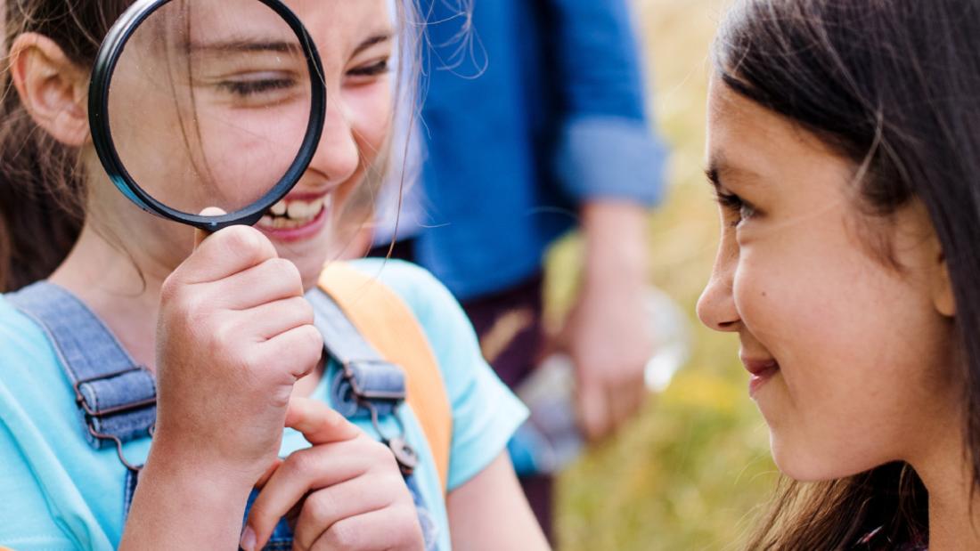 Two students smiling. One student takes notes as the other student looks through a magnifying glass outdoors.
