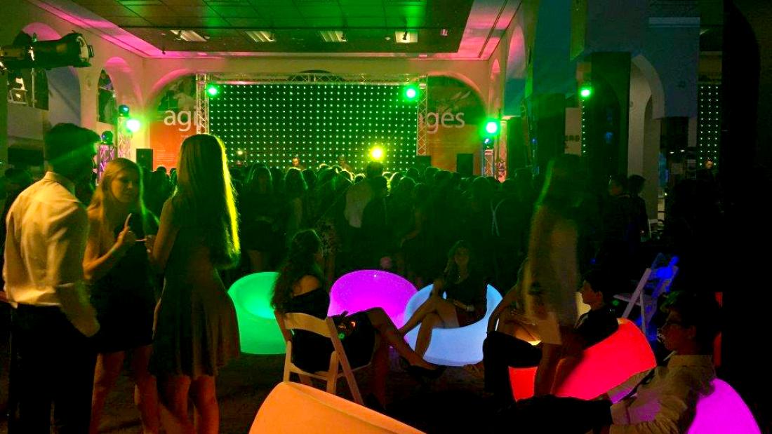 A room lit with neon green lighting. The room is crowded with the closest people talking while standing and while reclining in glowing orb chairs.