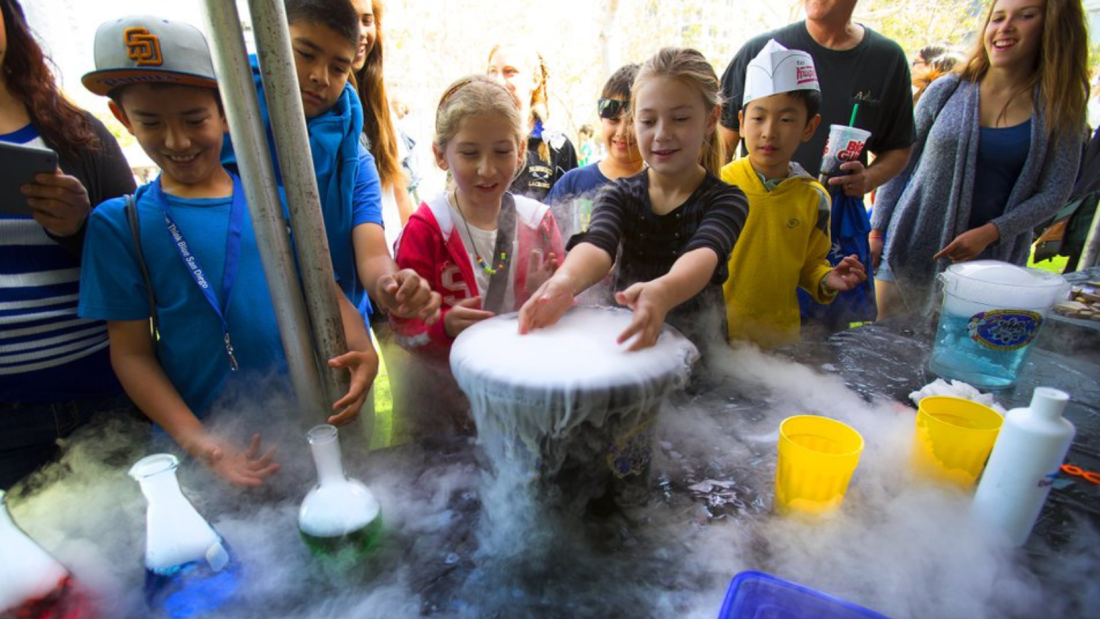 A group of eight students looking on and participating in a liquid nitrogen chemistry experiment in an outdoor setting. One student has their hands inside of a gray bowl overflowing with vapors.