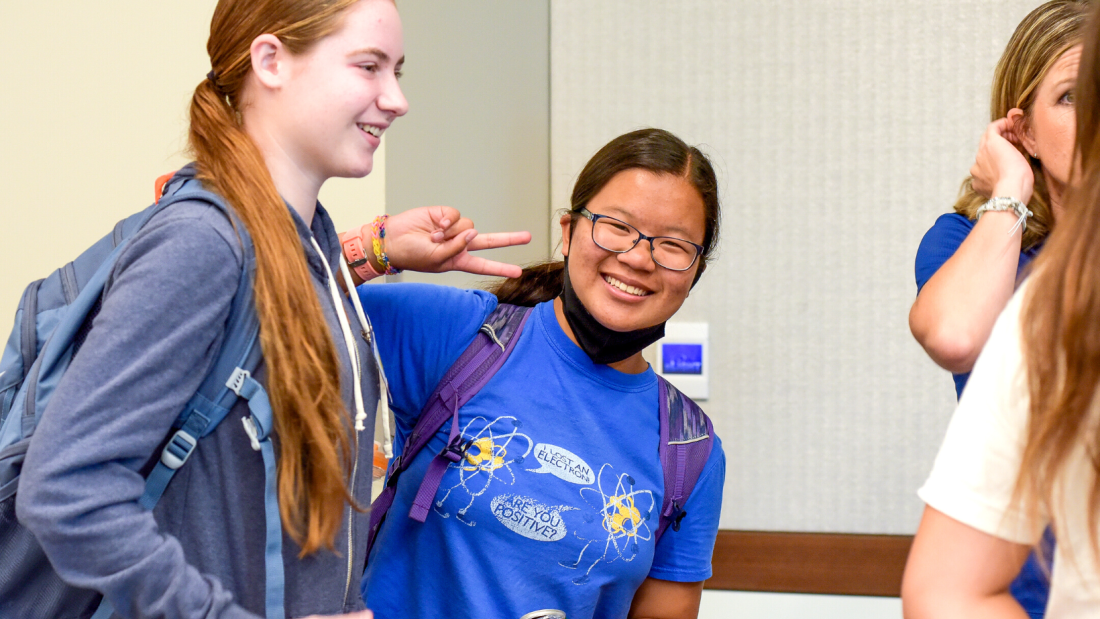 Two BEWiSE students at an event indoors. One student looks to the right of the frame as the other student is smiling at the camera holding up a peace sign.