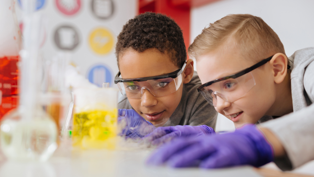 Two students focusing on a chemistry experiment inside of a lab. Both are wearing protective goggles and gloves.