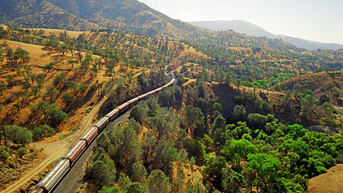 An aerial view of a long train cutting through a rolling hill landscape of green trees and yellow hillsides
