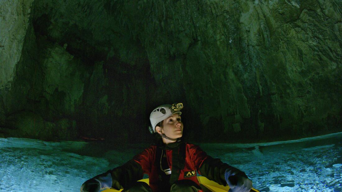 Scientist looking to their left inside of a large cave. The Sciectist is in a yellow raft atop blue water.