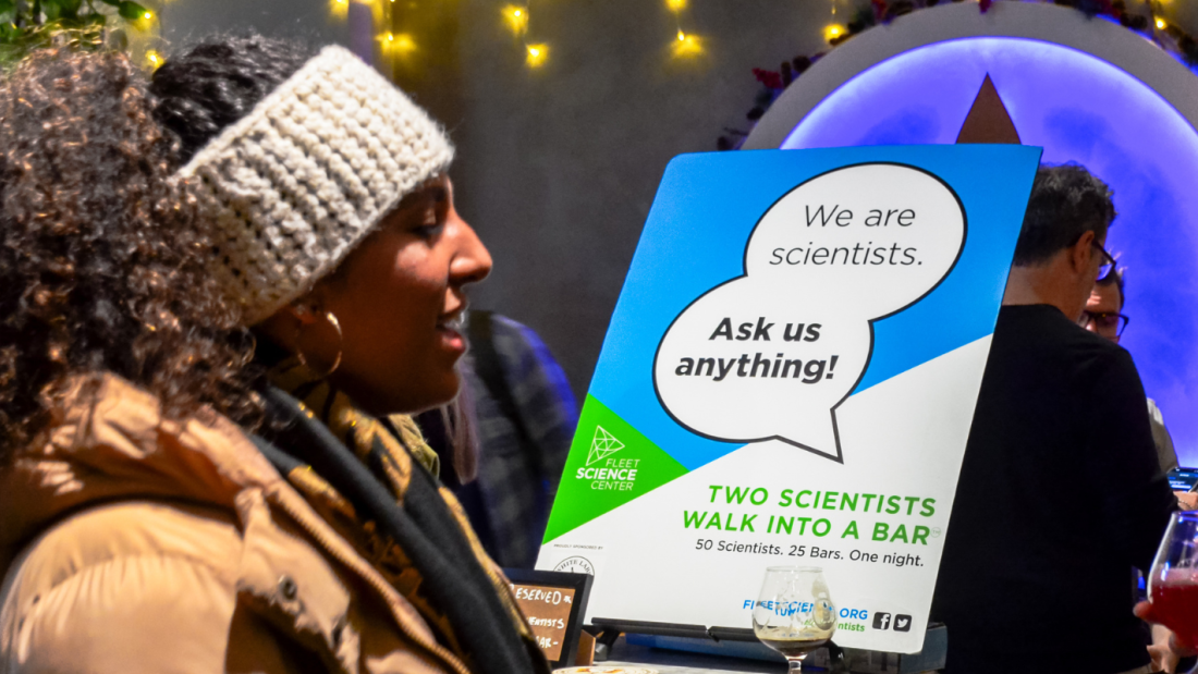 Woman stands in front of a sign that reads "We are scientists. Ask us anything!"