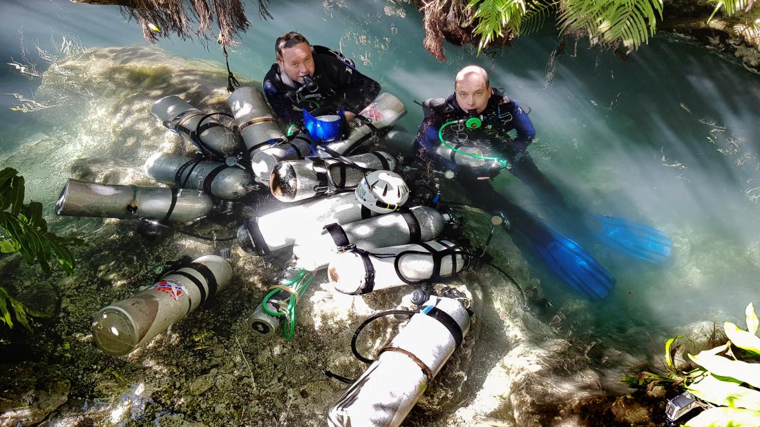 Two people in diving gear emerging from the water surrounded by oxygen tanks in an outdoor environment.