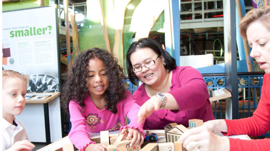 Two young girls with two women interact with building blocks on a table that look like houses.