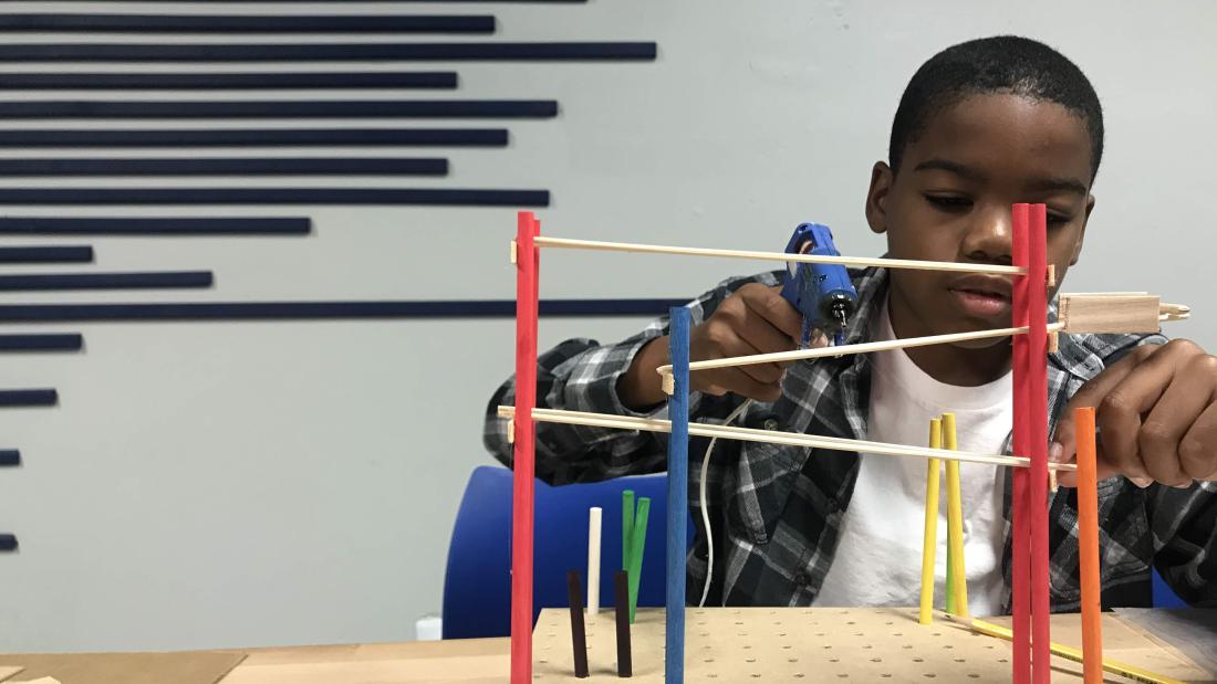 A black boy uses a hot glue gun on a vertical assembly of wooden dowels