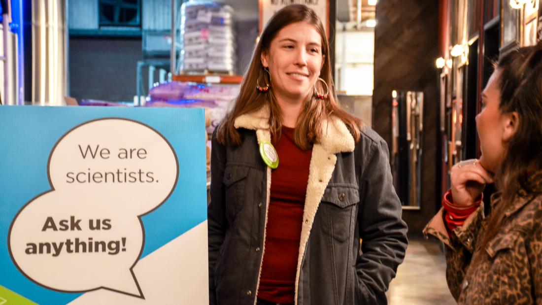 A woman in a sheepskin jacket stands near a sign that reads, "We are scientists. Ask us anything" at a brew pub. 