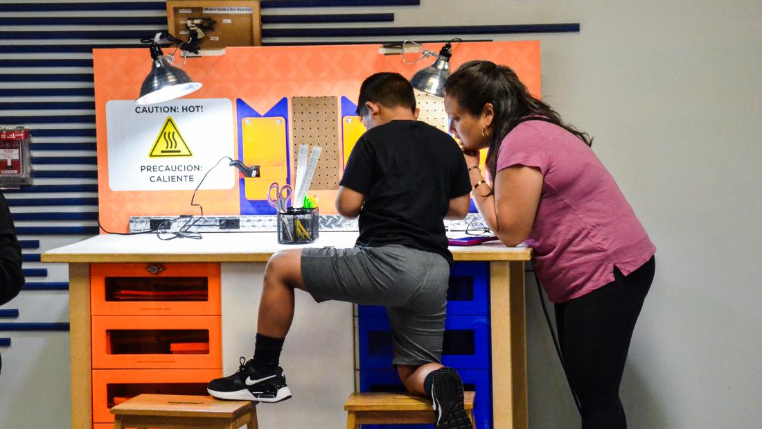 A boy kneels on a stool while a woman leans over to his side. They are building an unseen item on a work bench