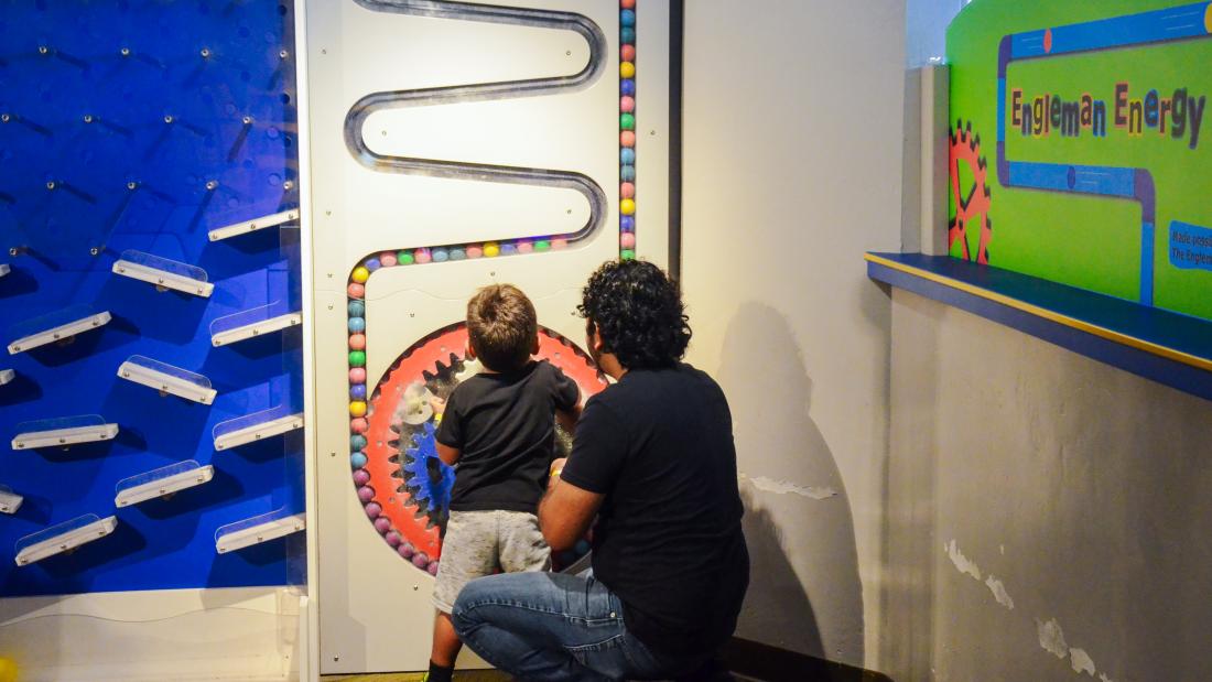 A man kneels down behind a young boy who is turning a large colorful wheel mounted into the wall. Curved tracks move up from the wheel to the ceiling out of sight