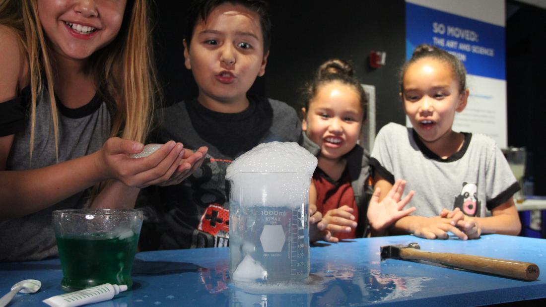 Three girl and a boy stand around a demo table with foaming beakers and look excited