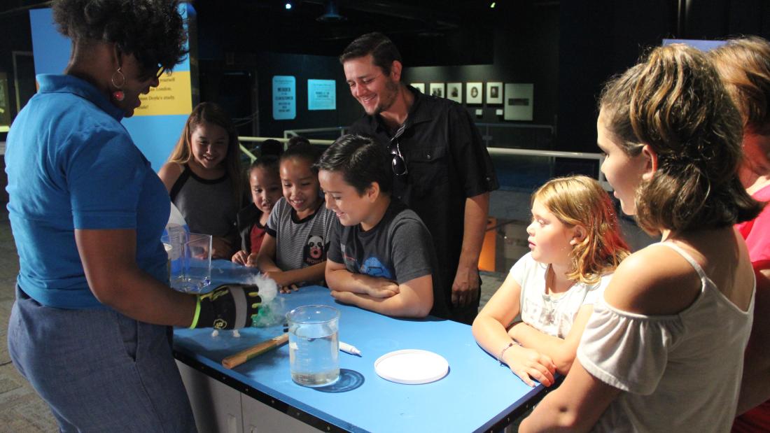 Adults and children stand around a blue demonstration cart while a woman wearing safety gloves demonstrates an experiment with beakers and liquid