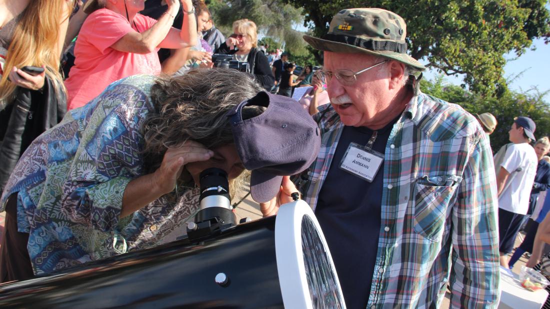 In a crowd a woman leans over an looks through a telescope while an older man watches over