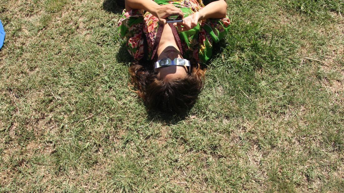 A woman lying on the grass upside-down so that her head is in the middle of the photo and her torso reaches off the top edge. She is wearing eclipse glasses and has her hands up