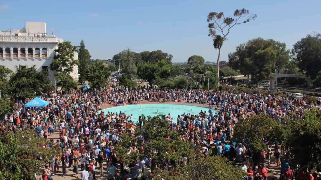 An aerial view of a very large crowd in Balboa Park surrounding a blue water fountain on a clear sky day.