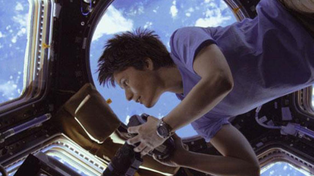 A woman floating from the upper right corner in a space station. She is holding a camera and the windows around her form a comb pattern. The earth is visible below