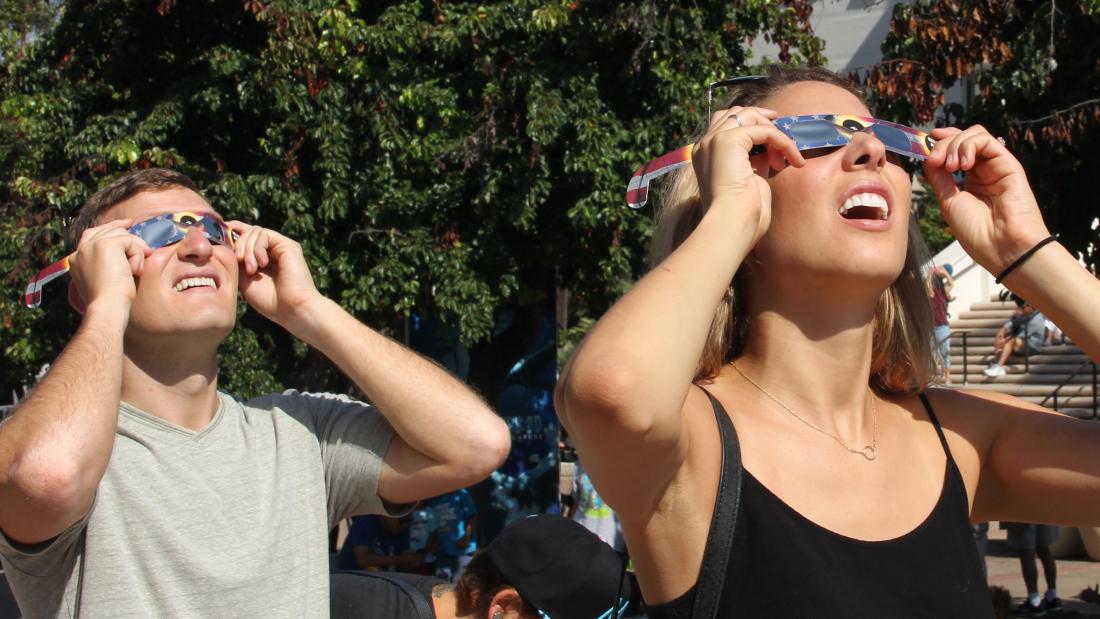 A man and woman wearing eclipse sunglasses squint up at the sun in a green outdoor area