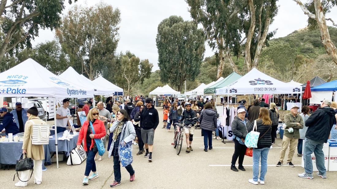 A crowded farmers market with white tents surrounded by large trees