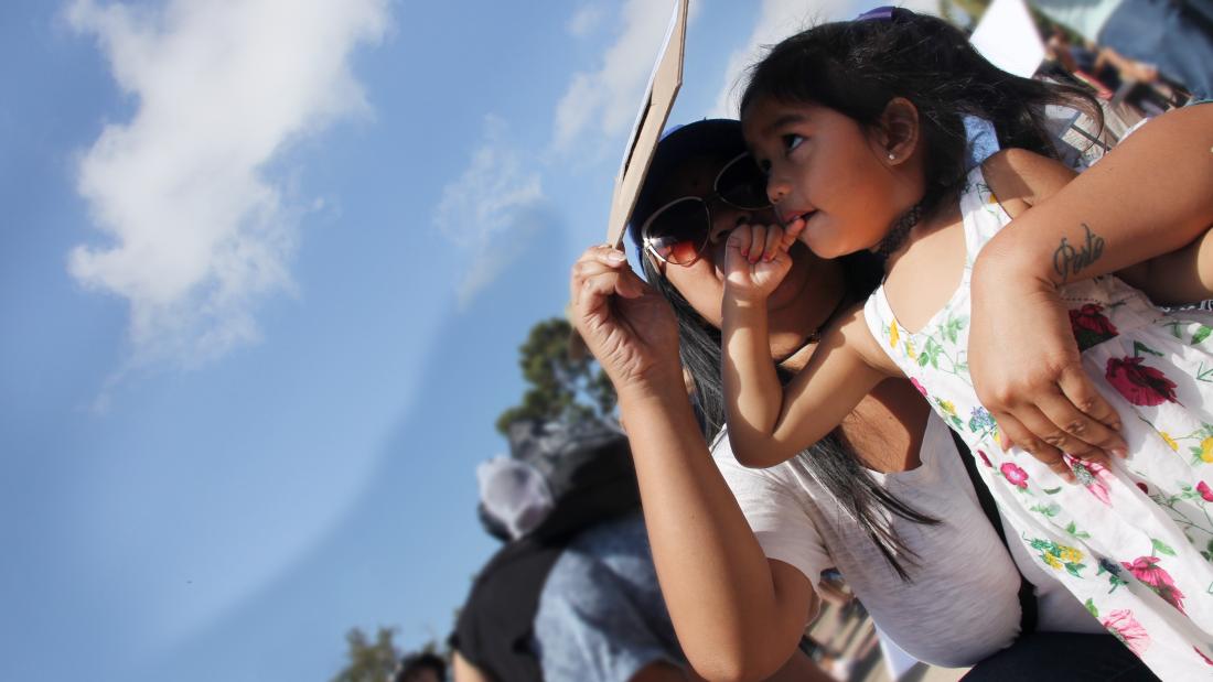 A woman holds a young girl and they both look up at the sky through an eclipse filter.