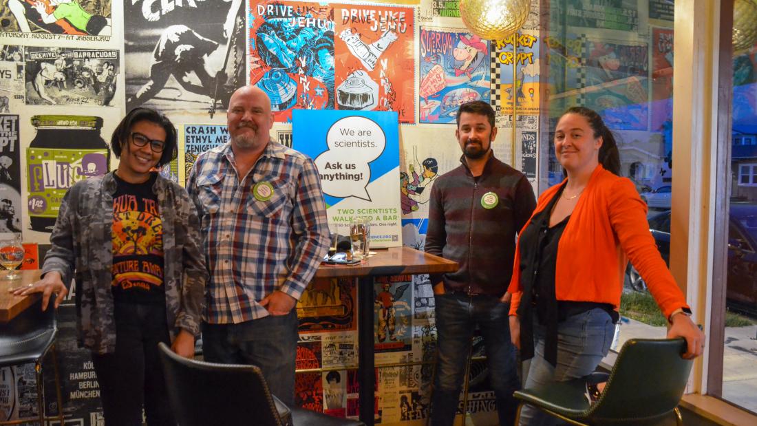 Four people standing around a bar high top table inside a colorful bar with a sign on the table.