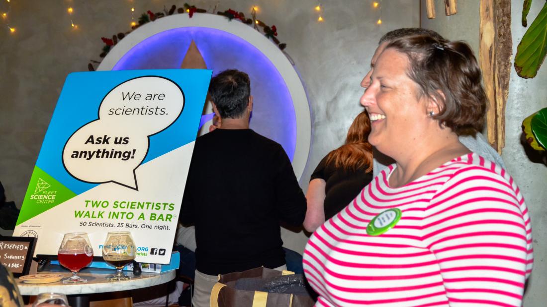 A woman laughs to the right of a sign on a table with beer glasses cluttered around