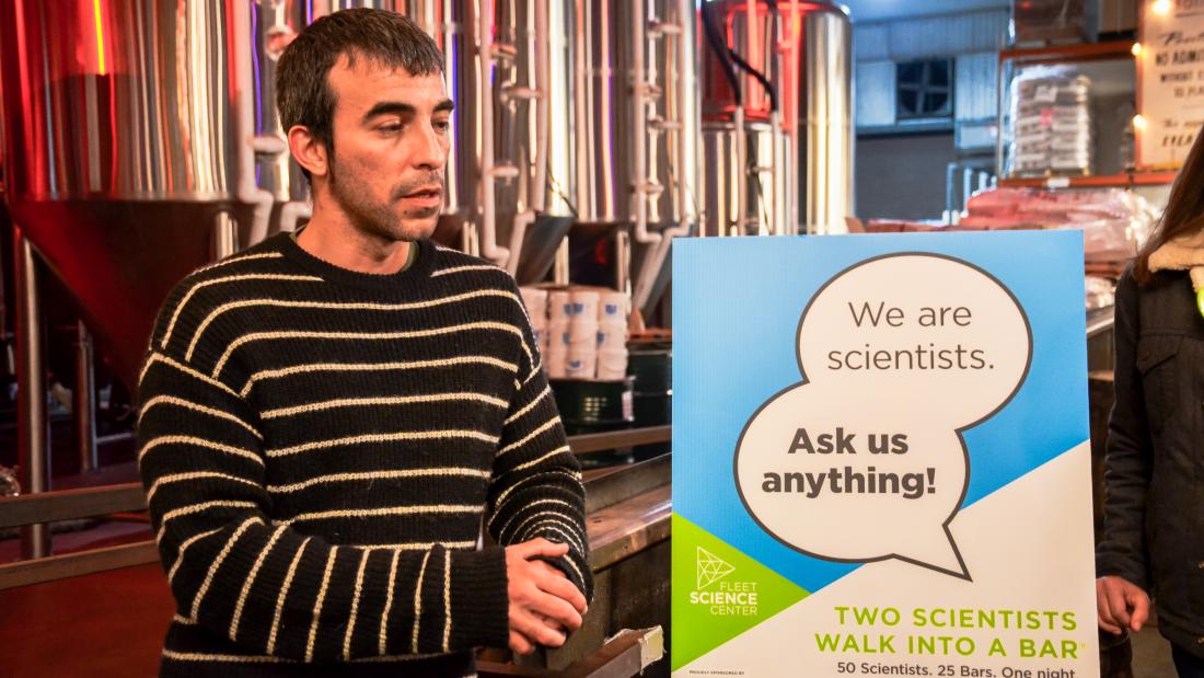 A man inside a brewery stands next to a sign that says We Are Scientists Ask Us Anything
