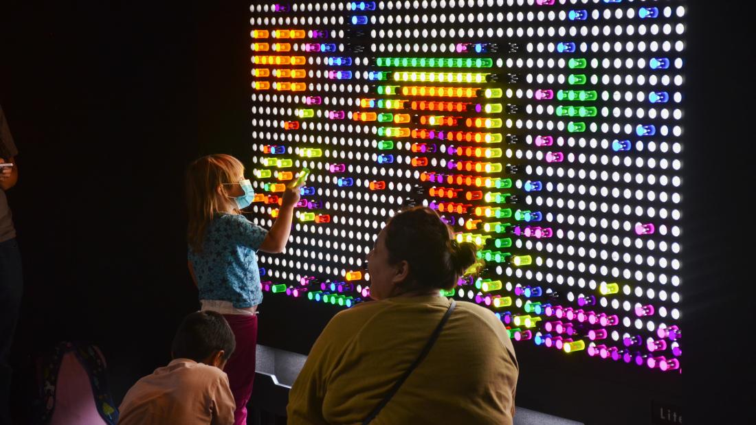 A woman sits on the floor watching a child play with a huge light brite in the dark