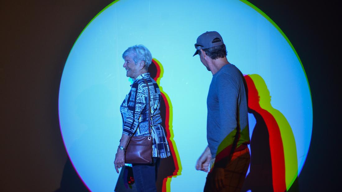 Two older adults walk past a lit circle on a wall. Their shadows are multiple colors.