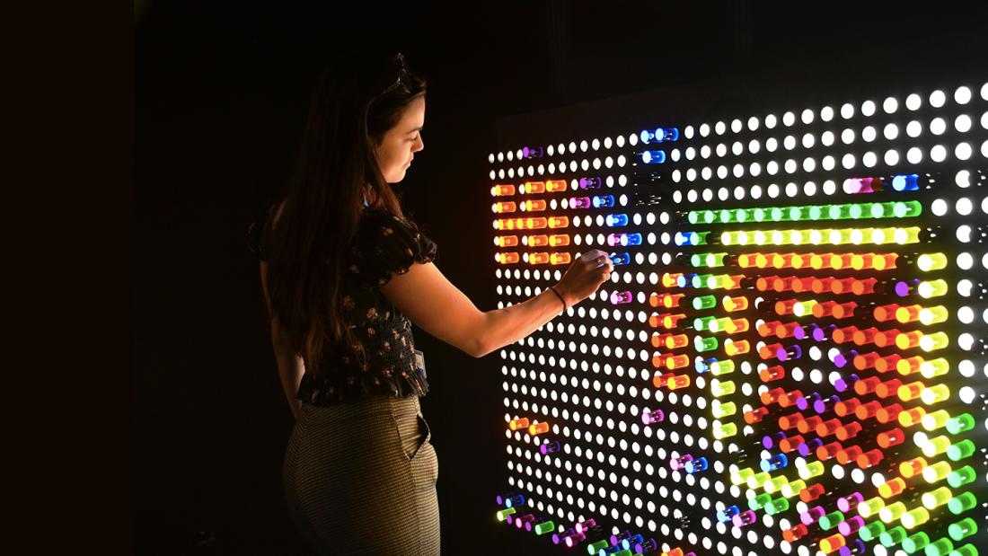 A woman standing in the dark playing with a huge lite brite on a wall