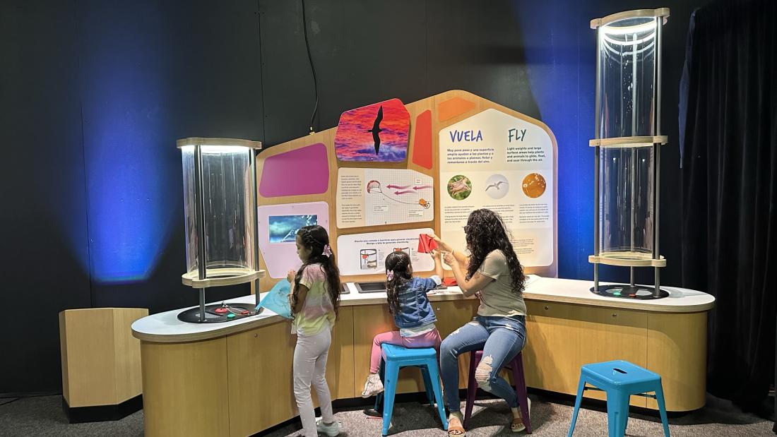 Two young girls and a woman sit together in front of a curved science exhibit in a darkened room