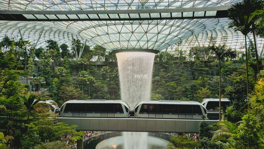 In this image, there is lots of vegetation in a room. In the center, there is a ceiling glass like a funnel and water pouring out. A train goes by.