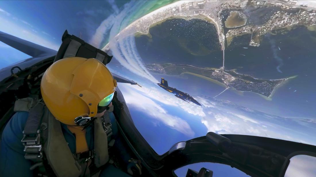 The image captures an exhilarating perspective from the cockpit of a Blue Angels jet during an aerial maneuver, seemingly captured during a training session or performance over a coastal area. The pilot, wearing a bright yellow helmet and reflective visor, can be seen in the foreground. The helmet is fastened securely, and various straps and buckles of the pilot's gear are visible.  Through the clear cockpit window, another Blue Angels jet is visible in the near distance, flying in formation. The jet is pai