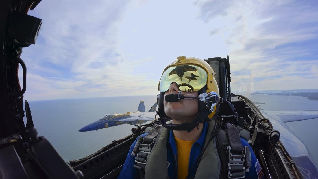 The image captures a mesmerizing view from the cockpit of a jet plane. Dominating the foreground is the left side of the cockpit, where the pilot is seen wearing a yellow helmet and a green-tinted visor, secured with multiple straps and buckles.  Beyond the cockpit, the exterior view is breathtaking. The sky transitions in hue from a clear blue to a mix of white and teal, indicative of the vast expanse of the ocean. Visible below, and seemingly inverted due to the plane's orientation, is a coastal landscape