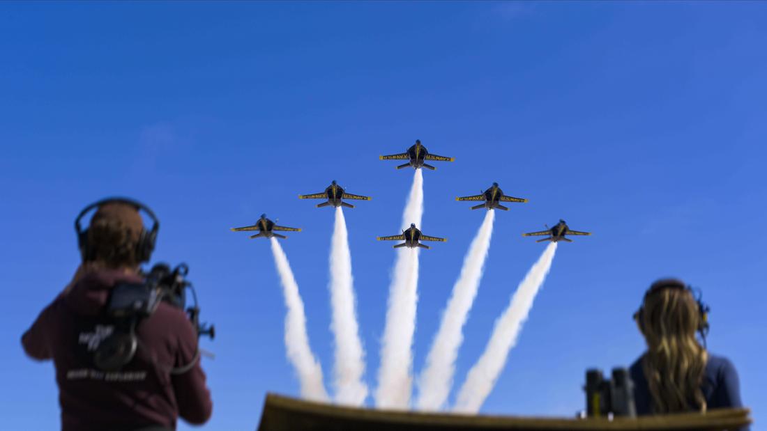 The image captures an airshow with six U.S. Navy Blue Angels jets soaring in formation against a bright, cloudless blue sky. The planes are positioned above the viewer, creating a dynamic sense of height and movement. Each jet leaves a white smoke trail, emphasizing their trajectory and adding visual excitement to the composition.  In the foreground, partially blurred and out of sharp focus, are two individuals with their backs to the camera. On the left is a person wearing headphones and a maroon-colored h