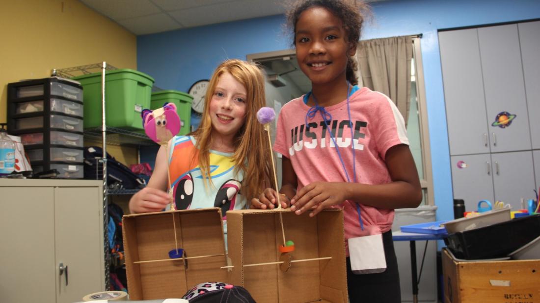 Two elementary-aged school girls smiling at the camera enjoying a science-based camp activity