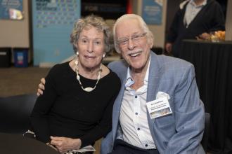 An older woman and man sit together with his arm around her and smile