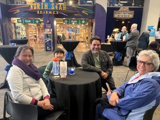 A woman, boy, man, and older woman sit around a small black round table and smile at the camera