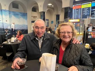 An older man and woman with his hand on her shoulder smile for the camera with drinks and a bag of popcorn on a hightop table in front of them