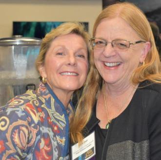 Two women lean their heads close together with the woman on the left smiling and the woman on the right laughing