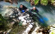 Two people in diving gear emerging from the water surrounded by oxygen tanks in an outdoor environment.