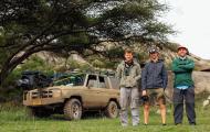 Three white men stand in front of a very muddy jeep under a tree