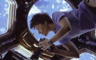 A woman floating from the upper right corner in a space station. She is holding a camera and the windows around her form a comb pattern. The earth is visible below