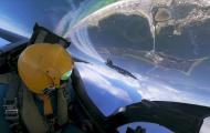 The image captures an exhilarating perspective from the cockpit of a Blue Angels jet during an aerial maneuver, seemingly captured during a training session or performance over a coastal area. The pilot, wearing a bright yellow helmet and reflective visor, can be seen in the foreground. The helmet is fastened securely, and various straps and buckles of the pilot's gear are visible.  Through the clear cockpit window, another Blue Angels jet is visible in the near distance, flying in formation. The jet is pai