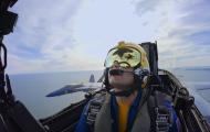 The image captures a mesmerizing view from the cockpit of a jet plane. Dominating the foreground is the left side of the cockpit, where the pilot is seen wearing a yellow helmet and a green-tinted visor, secured with multiple straps and buckles.  Beyond the cockpit, the exterior view is breathtaking. The sky transitions in hue from a clear blue to a mix of white and teal, indicative of the vast expanse of the ocean. Visible below, and seemingly inverted due to the plane's orientation, is a coastal landscape