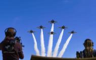 The image captures an airshow with six U.S. Navy Blue Angels jets soaring in formation against a bright, cloudless blue sky. The planes are positioned above the viewer, creating a dynamic sense of height and movement. Each jet leaves a white smoke trail, emphasizing their trajectory and adding visual excitement to the composition.  In the foreground, partially blurred and out of sharp focus, are two individuals with their backs to the camera. On the left is a person wearing headphones and a maroon-colored h