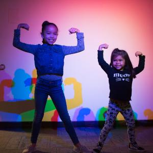 Two young girls stand in front of rainbow shadows making body builder poses and laughing