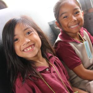 Two elementary school girls wearing maroon shirts smile at the camera while sitting on a school bus