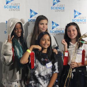Four girls pose with red award ribbons in front of a Fleet Science Center branded backdrop