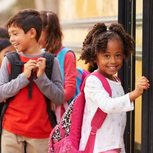 A young girl wearing a pink backpack walks onto a school bus and smiles at the camera