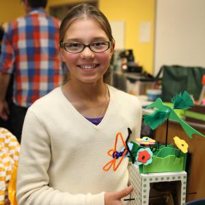 A young girl in glasses smiles at the camera while holding a craft made whimsical garden